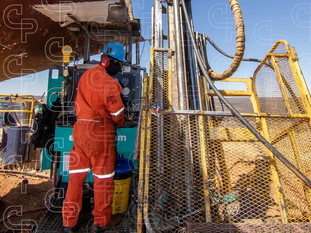 Person in orange protective clothing operating controls beside a drill rig with yellow guarding and hydraulic equipment.