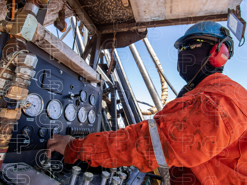 Person in protective workwear operating a drill rig control panel with gauges, levers and hydraulic lines.