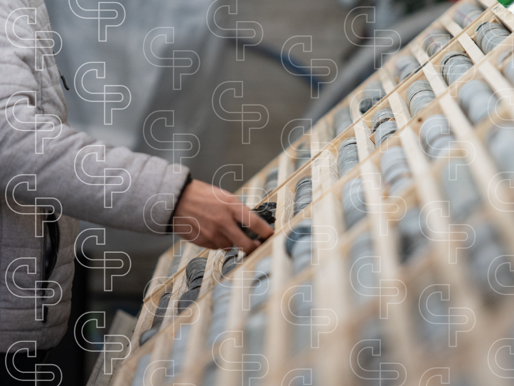 Person inspecting grey drill core samples arranged in a wooden core tray.