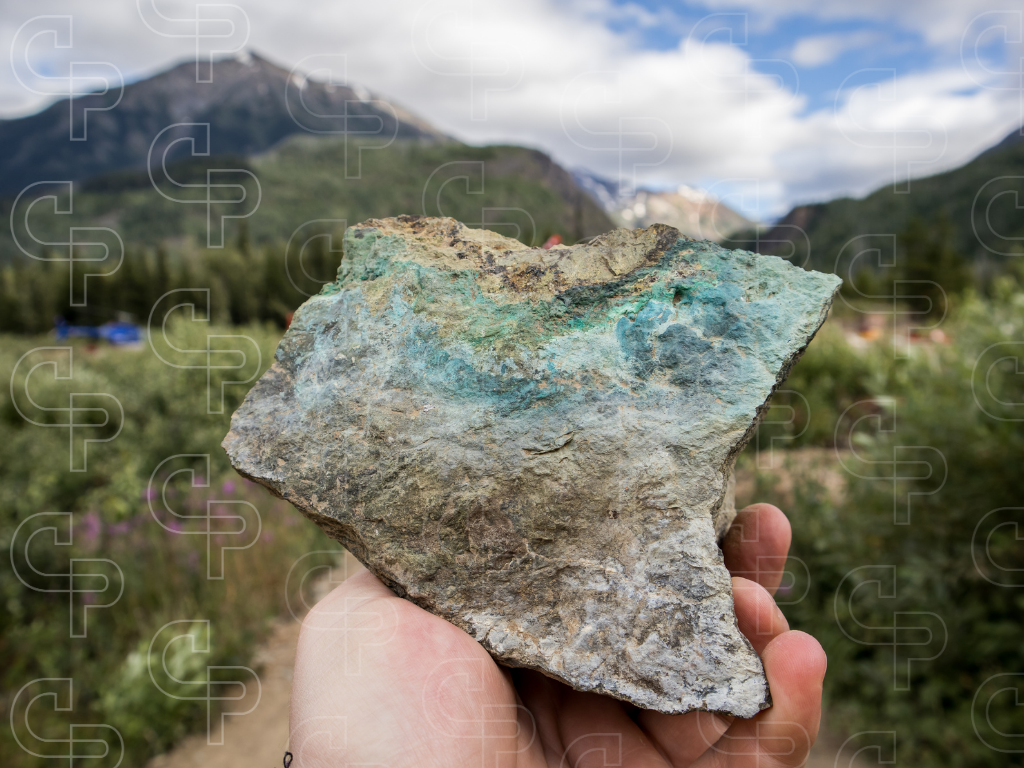 Hand holding a rock sample with blue-green copper mineralisation against a mountainous outdoor background.